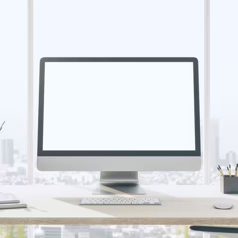 Frontansicht auf leeren weißen modernen Computermonitor auf hellem Holztisch mit Blumentopf, Tastatur und Notebooks im sonnigen Büro mit Stadtblick aus dem Fenster.