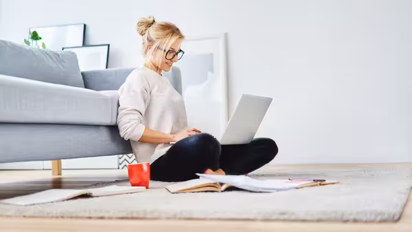 Foto: Eine junge Frau sitzt auf dem Teppich vor ihrer Couch und schreibt auf einem Notebook.