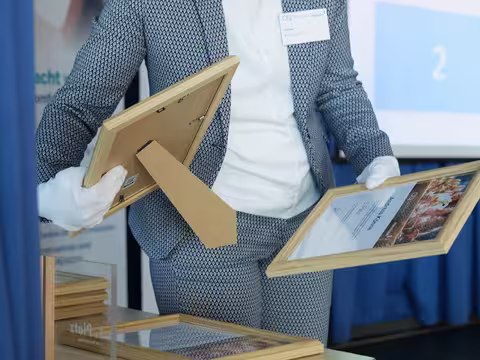 Foto: Nahaufnahme einer jungen Frau im schwarzweiß gemusterten Hosenanzug, die zwei der gerahmte Urkunden in ihre behandschuhten Hände nimmt.