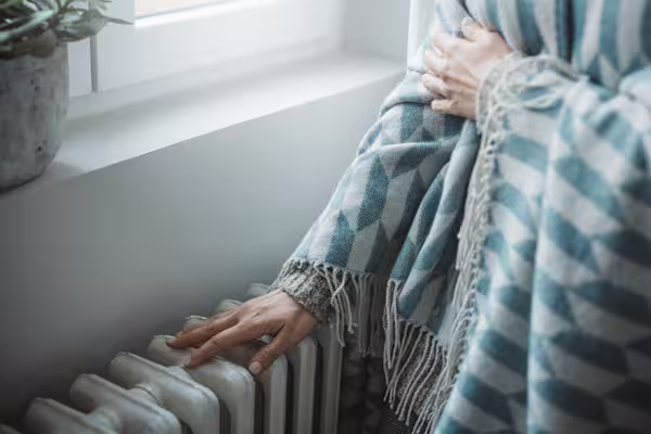 Person steht eingehüllt in eine Decke an einem Heizkörper und legt eine Hand auf die Heizkörperrippen.