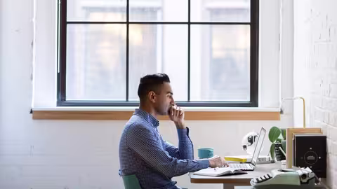 Person sitzt an einem Schreibtisch vor einem Laptop und arbeitet konzentriert in einem hellen Raum mit großem Fenster.