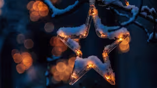 Foto: Ein fünfzackiger Stern aus durchsichtigem Glas hängt am schneebedeckten Zweig eines Baums, im dunkelblauen Hintergrund leuchten unscharfe LED-Lämpchen.