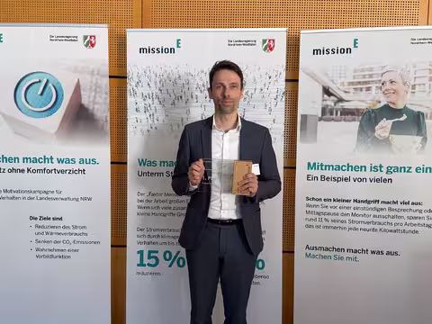 Foto: Mirco Schwettmann, ein junger schlanker Mann im dunkelblauen Anzug, vom Finanzamt Lübbecke hält die Auszeichnung für seine Behörden in die Kamera.