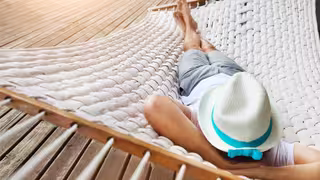 Eine Person liegt entspannt in einer weißen Hängematte aus geflochtenem Stoff. Sie trägt einen weißen Sonnenhut mit einem türkisfarbenen Band, ein weißes T-Shirt und graue Shorts. Die Person liegt auf dem Rücken, die Arme hinter dem Kopf verschränkt und die Beine übereinandergeschlagen.