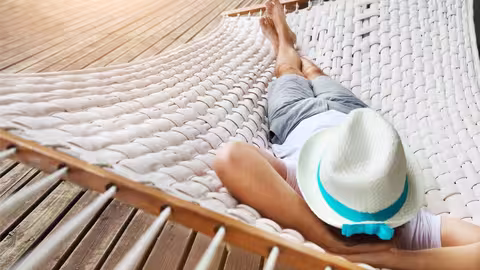 Eine Person liegt entspannt in einer weißen Hängematte aus geflochtenem Stoff. Sie trägt einen weißen Sonnenhut mit einem türkisfarbenen Band, ein weißes T-Shirt und graue Shorts. Die Person liegt auf dem Rücken, die Arme hinter dem Kopf verschränkt und die Beine übereinandergeschlagen.