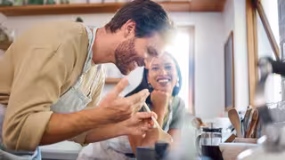 Zwei Personen stehen in einer Küche und kochen gemeinsam; eine Person rührt lachend in einem Topf, die andere schaut lächelnd zu.