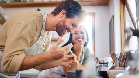 Zwei Personen stehen in einer Küche und kochen gemeinsam; eine Person rührt lachend in einem Topf, die andere schaut lächelnd zu.