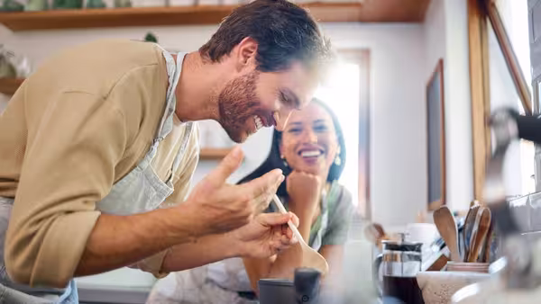 Zwei Personen stehen in einer Küche und kochen gemeinsam; eine Person rührt lachend in einem Topf, die andere schaut lächelnd zu.