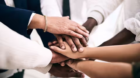 Foto: Eine Gruppe von Menschen steht im Kreis, die Hände in der Kreismitte übereinandergelegt.