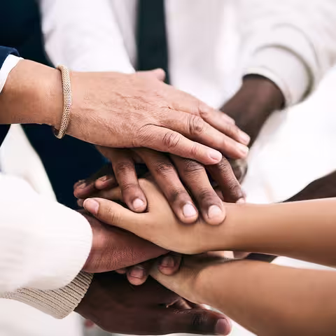 Foto: Eine Gruppe von Menschen steht im Kreis, die Hände in der Kreismitte übereinandergelegt.