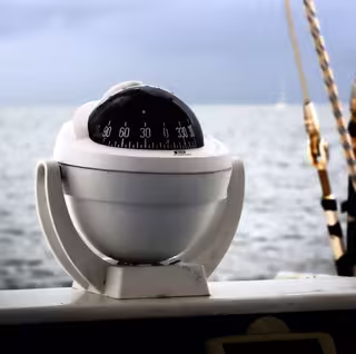 Foto: ein weißer Kugelkompass eines Schiffes, im Hintergrund das Meer.