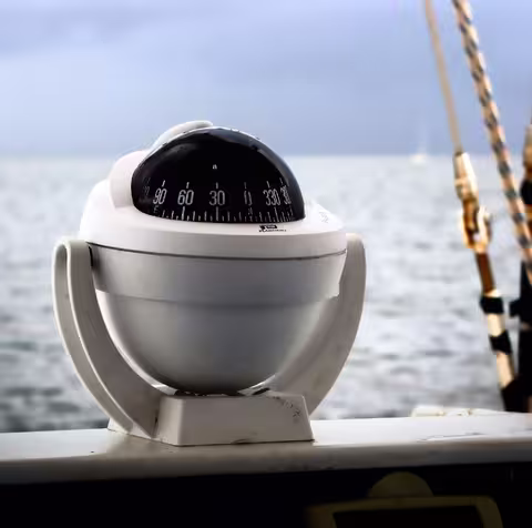 Foto: ein weißer Kugelkompass eines Schiffes, im Hintergrund das Meer.