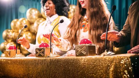 Foto: Retro-Gameshow mit goldfarbenen Luftballons, goldener Tischdecke und großen goldenen Buzzern von drei Kandidatinnen und Kandidaten