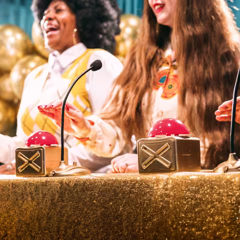 Foto: Retro-Gameshow mit goldfarbenen Luftballons, goldener Tischdecke und großen goldenen Buzzern von drei Kandidatinnen und Kandidaten