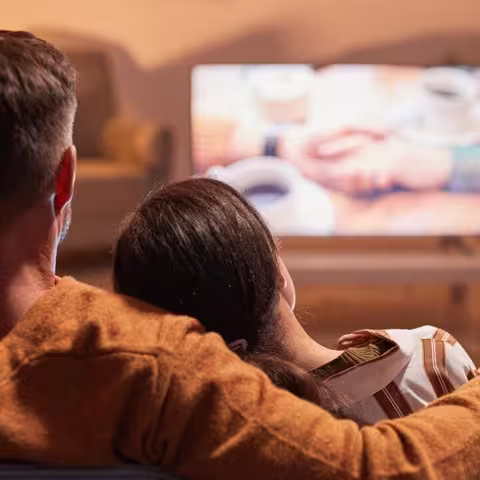 Rückansicht eines erwachsenen Paares, das zu Hause fernsieht, während es auf einem Sofa sitzt, das von warmem, gemütlichem Licht beleuchtet wird.