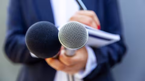 Foto: Nahaufnahme einer Journalistin oder Reporterin, die zwei Mikros in Richtung der Kamera und in der anderen Hand einen Notizblock hält