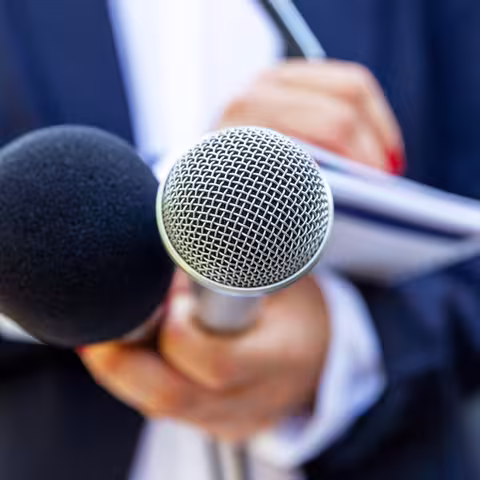 Foto: Nahaufnahme einer Journalistin oder Reporterin, die zwei Mikros in Richtung der Kamera und in der anderen Hand einen Notizblock hält