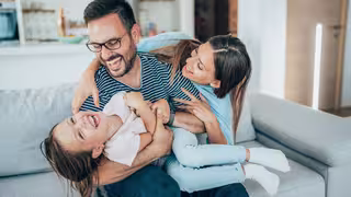 Foto: Zwei junge Eltern albern mit ihrer Tochter auf der Couch herum.