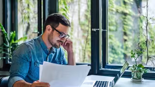 Person sitzt an einem Schreibtisch am Laptop und betrachtet Unterlagen, im Hintergrund große Fenster mit Pflanzen.