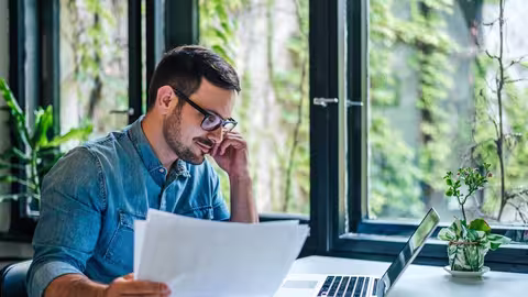 Person sitzt an einem Schreibtisch am Laptop und betrachtet Unterlagen, im Hintergrund große Fenster mit Pflanzen.