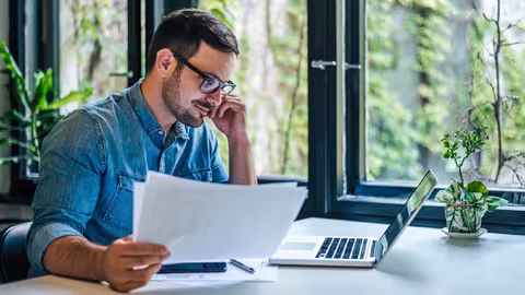 Foto: Ein junger Mann mit Brille und Dreitagebart hält ein paar Blätter Papier in der Hand und liest auf dem Display seines Laptops.