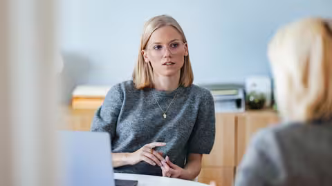 Foto aus der Perspektive der geöffneten Bürotür: Zwei Frauen unterhalten sich - die eine sitzt an ihrem Schreibtisch, die andere steht davor.