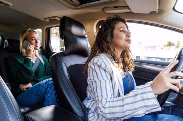 Fahrerin steuert ein Auto, während eine Frau auf dem Rücksitz telefoniert.