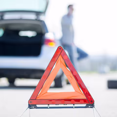 Foto: Ein weißer Pkw mit geöffnetem Kofferraum steht mit einer Panne am linken Straßenrand, dahinter steht – im Bildvordergrund – das rote Warndreieck.