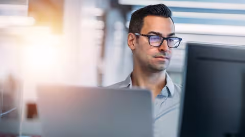 Foto: Ein junger Mann mit dunkler Brille sitzt in einem Büro an zwei Monitoren und arbeitet konzentriert am Computer.