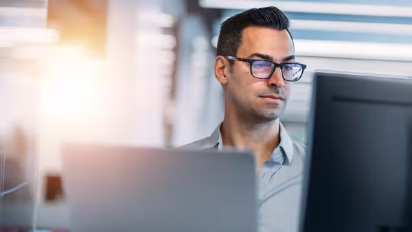 Foto: Ein junger Mann mit dunkler Brille sitzt in einem Büro an zwei Monitoren und arbeitet konzentriert am Computer.