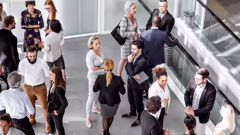 Foto: Mehrere Geschäftsleute stehen in kleinen Gruppen in einem offenen Bereich eines Gebäudes und reden miteinander.