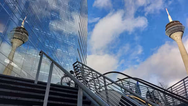 Auf dem Foto ist eine moderne Außentreppe mit einem Metallgeländer zu sehen, die zu einem Gebäudeeingang führt. Rechts im Bild ragt ein hoher Fernsehturm mit rot-weißer Spitze in den blauen Himmel. Links spiegelt sich derselbe Turm in der Glasfassade eines angrenzenden Gebäudes. Der Himmel ist teils bewölkt, teils blau. Die Perspektive ist leicht nach oben gerichtet, sodass die Architektur und der Turm imposant wirken.