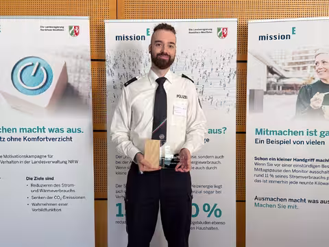 Foto: Till Hülsmann, ein groß gewachsener junger Mann mit dunklem Vollbart, hält seine Auszeichnung in die Kamera.