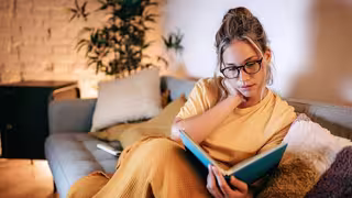 Person sitzt auf einem Sofa und liest ein Buch, eingehüllt in eine Decke in einer warm beleuchteten Wohnung.
