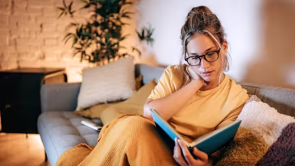 Person sitzt auf einem Sofa und liest ein Buch, eingehüllt in eine Decke in einer warm beleuchteten Wohnung.
