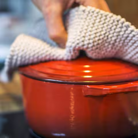 Foto: Jemand fasst mit einem dicken weißen Topflappen den Deckel eines roten Kochtopfs an, der auf dem Herd steht.