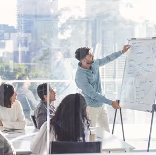 Foto: Geschäftsleute, die sich eine Präsentation auf dem Whiteboard ansehen. Ein Mann schreibt auf das Whiteboard mit Diagrammen und Grafiken, die anderen sitzen an einem Tisch davor. Alle sind leger gekleidet. Hinter ihnen befindet sich ein Fenster mit Blick auf die Stadt.