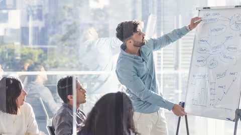 Foto: Geschäftsleute, die sich eine Präsentation auf dem Whiteboard ansehen. Ein Mann schreibt auf das Whiteboard mit Diagrammen und Grafiken, die anderen sitzen an einem Tisch davor. Alle sind leger gekleidet. Hinter ihnen befindet sich ein Fenster mit Blick auf die Stadt.
