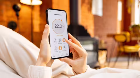 Foto: Eine Frau hält ein Handy mit laufender Smart-Home-Anwendung zur Heizungstemperaturregelung in der Hand, während sie entspannt auf der Couch liegt.