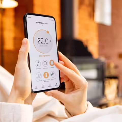 Foto: Eine Frau hält ein Handy mit laufender Smart-Home-Anwendung zur Heizungstemperaturregelung in der Hand, während sie entspannt auf der Couch liegt.