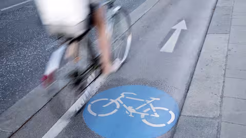 Foto: Teilansicht einer Radfahrerin auf einem Radweg neben einer Fahrbahn, seitlich von hinten fotografiert