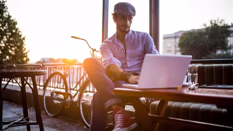 Foto: Ein leger gekleideter junger Mann mit Schirmmütze sitzt neben seinem Fahrrad an einem Tisch im Freien und arbeitet an seinem Notebook.