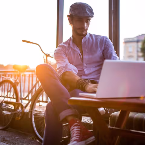 Foto: Ein leger gekleideter junger Mann mit Schirmmütze sitzt neben seinem Fahrrad an einem Tisch im Freien und arbeitet an seinem Notebook.