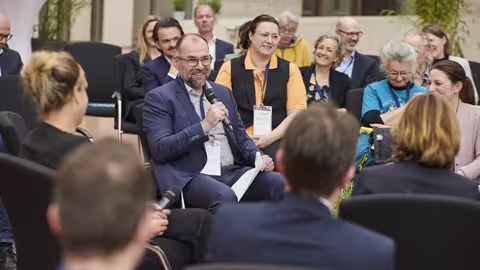 Foto: In der Mitte des in mehreren Kreisen sitzenden, lächelnden Publikums diskutiert eine kleine Gruppe von Vertreter:innen aus verschiedenen Behörden der Landesverwaltung NRW.