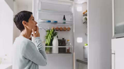 Eine Frau mit kurzen schwarzen Haaren steht vor einem geöffneten Kühlschrank und wirft einen prüfenden Blick auf die Lebensmittel.