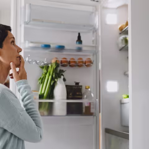 Eine Frau mit kurzen schwarzen Haaren steht vor einem geöffneten Kühlschrank und wirft einen prüfenden Blick auf die Lebensmittel.