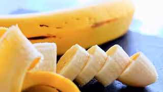 Auf dem Bild liegt eine geschälte Banane auf einer dunklen Oberfläche. Ein Teil der Banane ist in gleichmäßige Scheiben geschnitten, die schräg nebeneinanderliegen. Im Hintergrund befindet sich der ungeschälte Rest der Banane. Das Bild ist hell ausgeleuchtet und zeigt die Textur und Farbe der Frucht deutlich.
