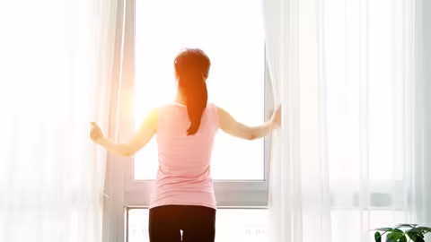 Eine Person steht in einem hellen Raum vor einem großen Fenster. Sie hält mit beiden Händen die weißen Vorhänge zur Seite und blickt nach draußen ins Sonnenlicht. Die Szene wirkt ruhig und frisch, das Licht fällt sanft in den Raum. Links und rechts des Fensters hängen leichte, helle Gardinen. Rechts im Bild ist eine grüne Zimmerpflanze zu sehen.