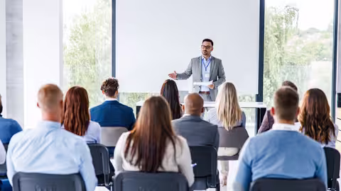 Foto: Blick von hinten in einen Tagungsraum, wo vor ein Mann zu seinem Publikum spricht