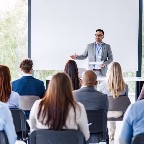 Foto: Blick von hinten in einen Tagungsraum, wo vor ein Mann zu seinem Publikum spricht